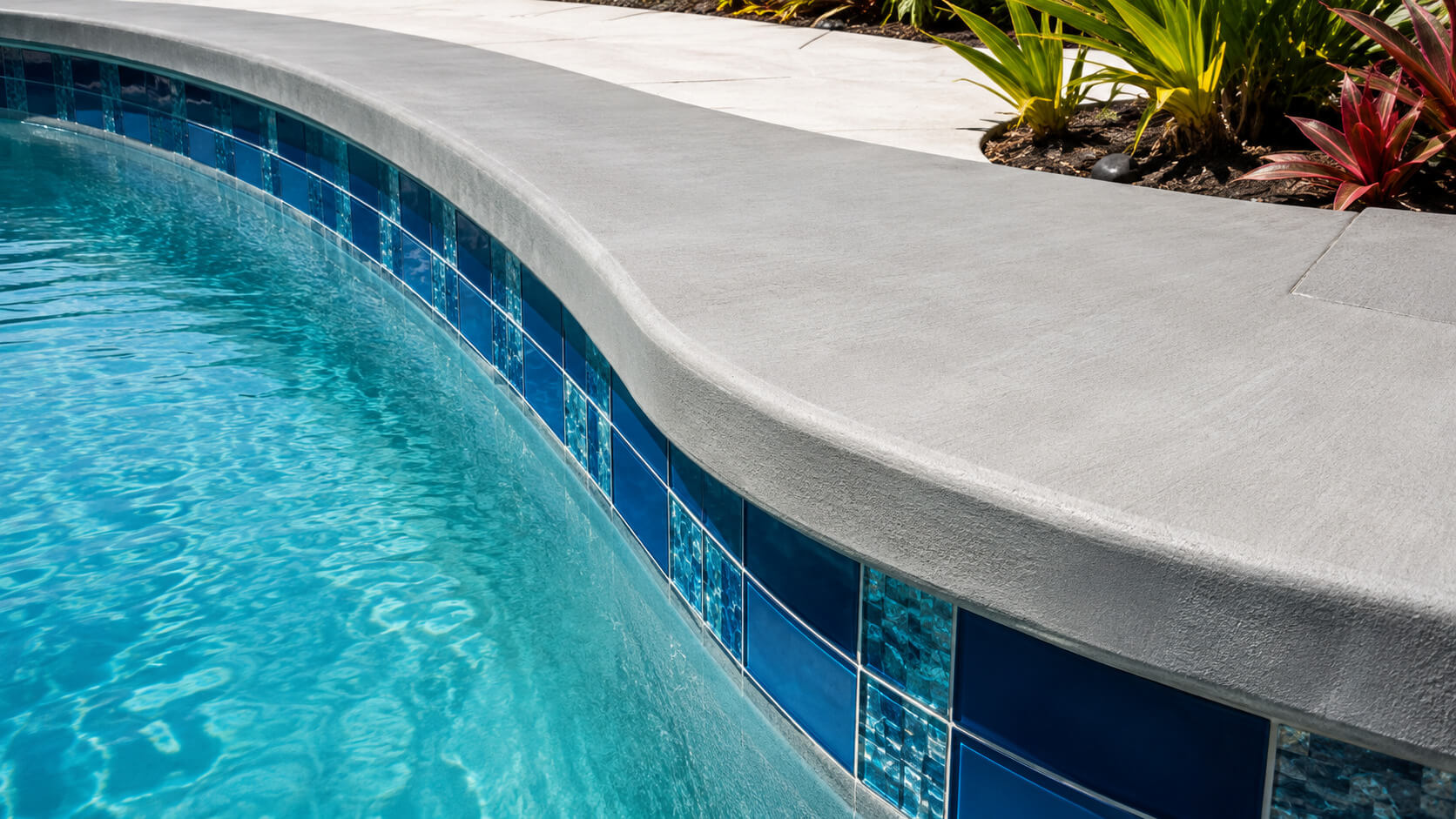 Gray brushed poured concrete coping on a curved pool edge above blue glass waterline tile