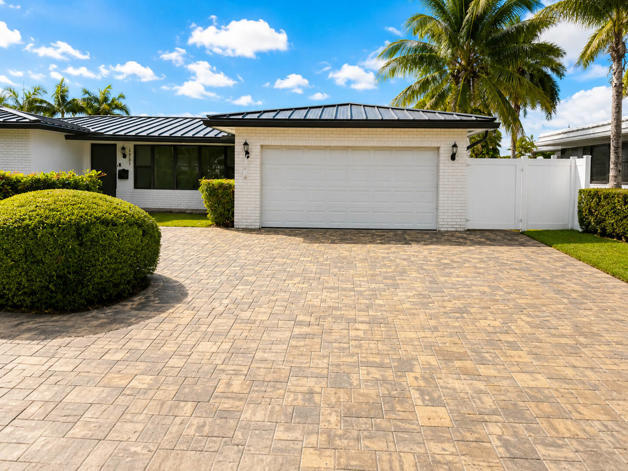 Beige running bond paver driveway at a white brick home with a black standing seam metal roof in Gulfstream