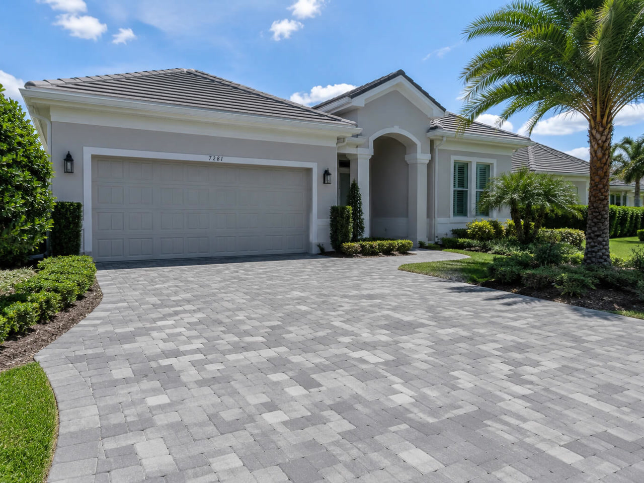 Gray tumbled paver driveway curving toward the two car garage of a Gulfstream home, framed by clipped hedges and palms