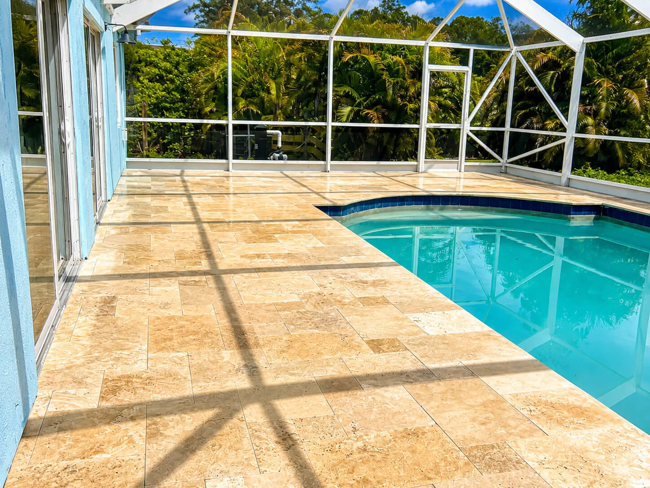 Sand toned travertine paver pool deck under a white screen enclosure on a Jupiter property, with dark blue tile coping along the pool