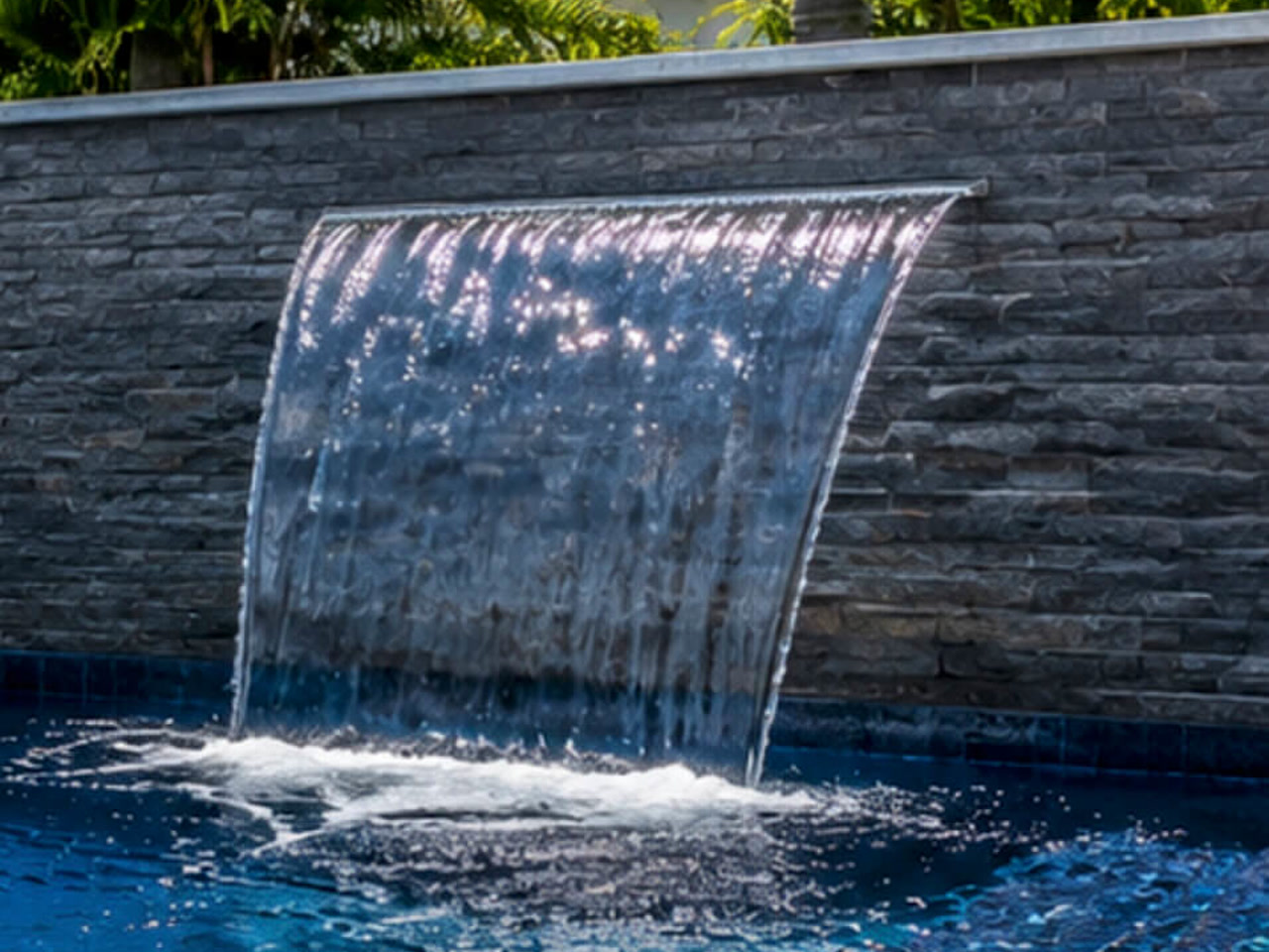 Sheer descent waterfall blade falling from a gray stacked stone wall into a deep blue pool at a Gulfstream estate