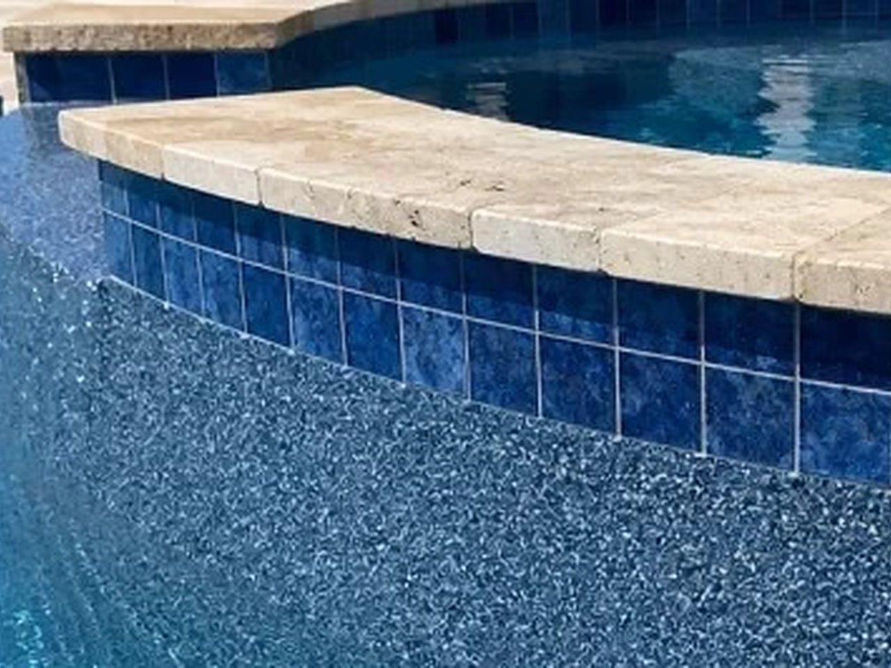 Blue pebble pool finish meeting a raised spa wall of deep blue tile capped with travertine coping