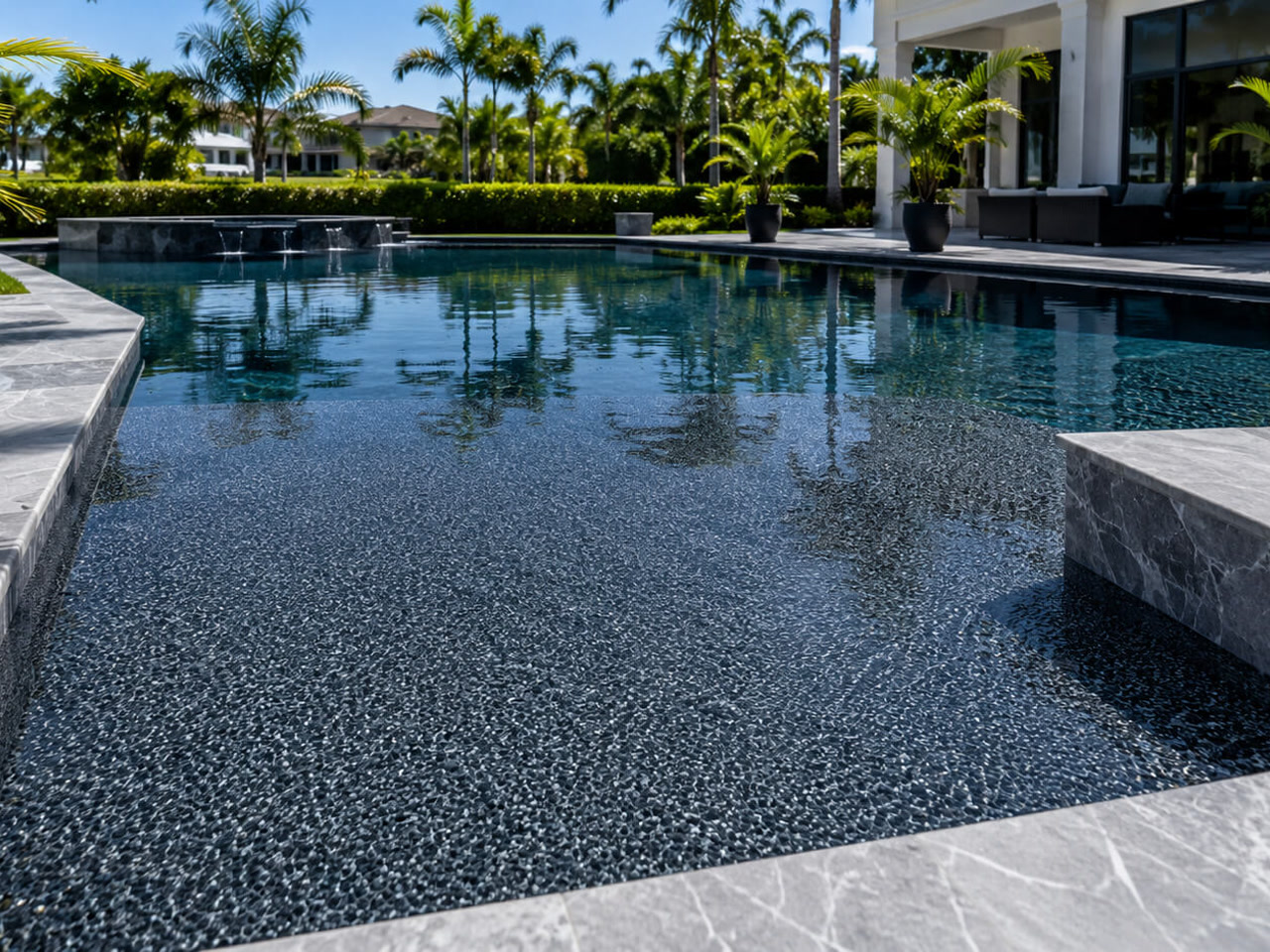 Dark charcoal pebble pool finish under still water, framed by gray marble coping and spillway