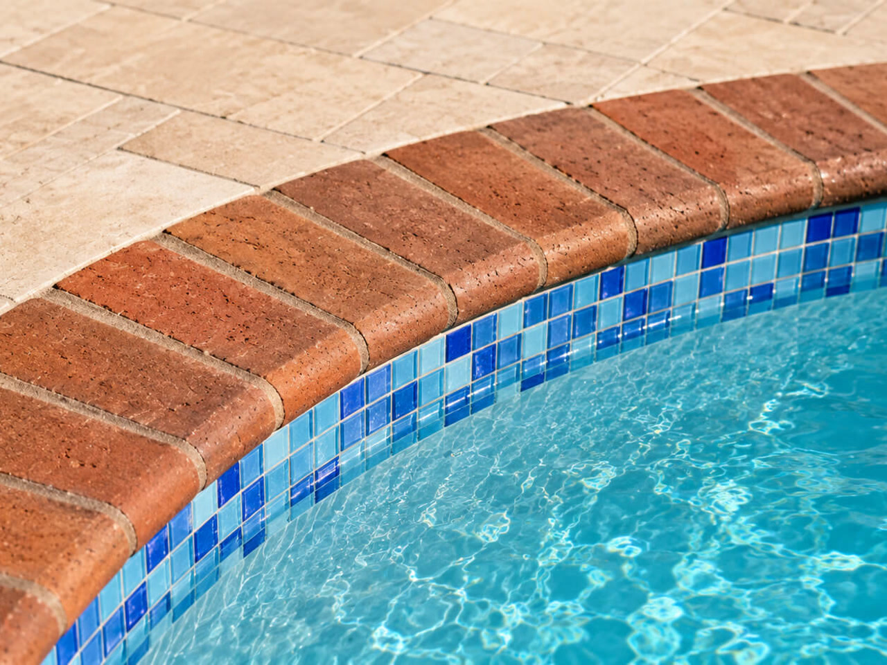 Red brick coping following the curve of a pool over a blue mosaic waterline tile, beside a travertine deck